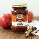 A jar of Old School Brand Apple Butter sits on a white surface beside two red apples, cinnamon sticks, and cloves. The orange and white label shows this 16 oz (510 g) old fashioned spread from Old School Brand.