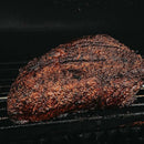 A large, seasoned beef brisket with a dark, crispy bark cooks on the grill, its rich flavors brought out by Dead Rooster Co. Old Timers All-Purpose Dry Rub. Distinct grill marks are visible on the tender meat, set against a dark background.