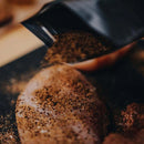A close-up of Dead Rooster Co Old Timers All-Purpose Dry Rub being poured over raw chicken breasts from Dead Rooster Co., seasoning them on a dark surface in preparation for cooking.