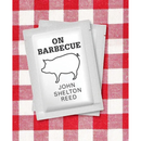 A white packet featuring a pig illustration and the title "On Barbecue – A Thought-Provoking Look at America's Barbecue Culture" by Books by splitShops rests on a red and white checkered tablecloth, perfectly capturing the rich tradition and flavors of American barbecue. This engaging BBQ book invites readers to explore the history and culture of barbecue through its charming design.