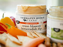 A close-up of Terrapin Ridge Farms Onion Blossom Horseradish Dip in a jar, surrounded by vibrant bell peppers and crackers, showcases this savory Terrapin Ridge Farms dipping sauce. Another jar of the same dip is partially visible beside it.