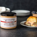 A jar of Terrapin Ridge Farms Onion Blossom Horseradish Dip sits on a dark surface beside a sandwich with meat, melted cheese, and sauce. Blurred stacked plates appear in the background.