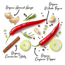 An assortment of spices and herbs, including two red cayenne peppers, sliced onions, cinnamon sticks, ground ginger, thyme, garlic, rosemary, and peppercorns, is arranged on a white background with handwritten labels identifying each spice. These ingredients are featured as part of The Spice Lab Organic Spice Complete Collection – 30 Jars by The Spice Lab, showcasing USDA certified organic spices.