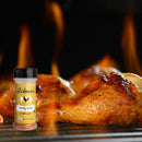 Juicy grilled chicken legs sizzle over open flames with a bottle of Hickman's BBQ Party Fowl Poultry Package from Hickman's BBQ in the foreground, highlighting a mouthwatering barbecue scene elevated by flavorful BBQ chicken seasoning.