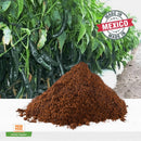 A pile of Rico Rico Pasilla Chile Powder Shaker, an essential ingredient in Mexican cuisine from the Rico Rico brand, is displayed in front of green pasilla chile plants. A "Made in Mexico" seal appears at the top right, and a small label at the bottom left reads "Pasilla Chile Powder.