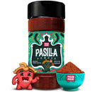 A bottle of Rico Rico Pasilla Chile Powder Shaker featuring a decorative label, accompanied by a small bowl of the rich red-brown spice and a lively red cartoon pepper with a green stem—ideal for infusing your favorite Mexican dishes with authentic Pasilla chile flavor from Rico Rico.