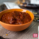 A rustic orange bowl brimming with rich, chunky red salsa sits on a wooden table, evoking the vibrant flavors of authentic Mexican cuisine. In the blurred background, ingredients—possibly including Rico Rico Pasilla Chile Powder Shaker—along with its packaging are visible. The image displays the “Rico Rico” logo in the bottom right corner.