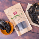 A package of Rico Rico Pasilla Dried Chiles – Mild & Rich Mexican Chile Negro from Oaxaca rests beside a small bowl and a napkin, each holding dried pasilla chiles, on a wooden surface—ideal for creating authentic Oaxacan mole negro recipes.