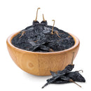 A wooden bowl filled with Rico Rico Pasilla Dried Chiles – Mild & Rich Mexican Chile Negro from Oaxaca, with two additional dried chiles from the Rico Rico brand in front of the bowl on a white background—essential ingredients for traditional mole negro.