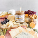 A wooden cheese board spread includes assorted cheeses, crackers, salami, blueberries, grapes, cashews, orange slices, pickles, and a jar of Terrapin Ridge Farms Terrapin Ridge Pecan Honey Mustard—a sweet and savory centerpiece.