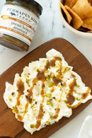 A wooden board with whipped cheese spread, drizzled with Terrapin Ridge Farms Pecan Honey Mustard and sprinkled with chopped pistachios. A jar of the mustard and a bowl of pita chips sit nearby.