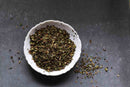 A white bowl filled with World Spice Peppermint from World Spice sits on a dark gray surface, with some of the dried, crushed herbs scattered around the bowl.
