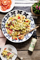 A large bowl of pasta salad with olives, cherry tomatoes, and cheese sits on a rustic table. Next to it is a Terrapin Ridge Farms Pesto Aioli Squeeze 12.5 oz bottle, sliced tomatoes, a plant, a napkin, and an aioli squeeze bottle.