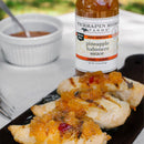 Grilled chicken breasts topped with Terrapin Ridge Farms Pineapple and Habanero Sauce are served on a dark wooden board, with a bottle of the sauce and a white ramekin filled with extra Terrapin Ridge Farms sauce in the background.