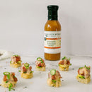 A bottle of Terrapin Ridge Farms Pineapple and Habanero Sauce is seen behind appetizers topped with sliced jalapeños and the sweet, spicy sauce from Terrapin Ridge Farms, all arranged on a white surface.