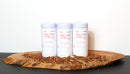 Three white canisters labeled World Spice Pink Chili Crisp from World Spice are arranged on a wooden tray, surrounded by scattered chili flakes and a small gold spoon, set against a white background. This distinctive spice blend features pink peppercorns for an unexpectedly vibrant twist.