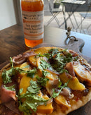 A flatbread topped with peach, prosciutto, greens, and sauce rests on a wooden board. Behind it sits a bottle of Terrapin Ridge Farms Hot Pepper Peach Bourbon Sauce by Terrapin Ridge Farms, with patio furniture visible in the background.