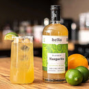 A bottle of Hella Cocktail Co. Classic Margarita Mix next to a tall glass filled with a yellow drink, garnished with a lime wedge and a salted rim, with two limes and an orange on a wooden surface.