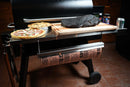 A Pimp My Grill® Tactical Stainless Folding Front Shelf for Traeger Pro 780 / Ironwood 885 holds a brisket on a wooden board, a pizza, and a pie, with a roll of butcher paper hanging from the shelf’s integrated rack.