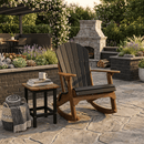The Kanyon Living Adirondack Rocking Chair Black & Deluxe Antique Mahogany sits on a stone patio beside a small table with a mug and book, a blanket and basket nearby, surrounded by lush plants and a stone outdoor fireplace.
