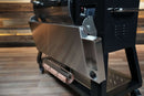 A stainless steel Pimp My Grill® Tactical Stainless Folding Front Shelf for the Recteq RT-800 / Deck Boss 800 attached to a pellet grill, featuring a side cut-out for tools and a paper towel holder underneath for a convenient prep workspace.