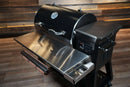 Black Recteq RT-800 / Deck Boss 800 pellet grill with a Pimp My Grill® Tactical Stainless Folding Front Shelf featuring a paper towel holder and integrated accessory tray as a prep workspace.