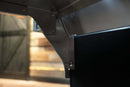 A metal bracket mounting a Pimp My Grill® Tactical Stainless Folding Front Shelf for Recteq RT-700 / RT-680 to the side of a barbecue.