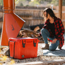 Red 25-quart cooler by Bison Coolers with black latches, sitting outside as a woman adds firewood to a chiminea.
