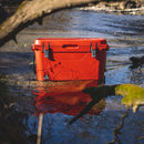 Red 50 quart cooler by Bison, sitting in shallow creek water with reflections and tree branches framing the shot.
