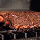 Close-up of a seasoned steak with a dark, peppery crust, coated in Dead Rooster Co Redwood BBQ Rub by Dead Rooster Co., cooking on a grill with grill grates visible underneath.