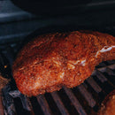 A seasoned brisket cooks on a grill, developing a crispy brown crust as it smokes over the grates, enhanced by Dead Rooster Co.’s Redwood BBQ Rub, which brings out deep, smoky flavors reminiscent of Kansas City barbecue.