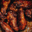 A close-up of several crispy, seasoned chicken wings showcases a dark, caramelized coating reminiscent of classic Kansas City barbecue. The wings appear freshly cooked, juicy, and flavorful, their slightly glossy surface and visible charred spices highlighting the signature Dead Rooster Co Redwood BBQ Rub from Dead Rooster Co.