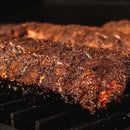 Close-up of seasoned and grilled racks of ribs resting on a grill, their crispy, dark crust revealing the bold flavors created by Dead Rooster Co Redwood BBQ Rub from Dead Rooster Co.