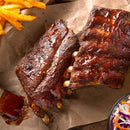 Two racks of barbecued ribs with Saltopia BBQ Salt: MEAT HEAD by SALTOPIA Infused Sea Salts rest on brown parchment, served with seasoned fries, coleslaw, and a nearby basting brush with sauce.