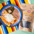 A plate of enchiladas topped with cheese, refried beans, and roasted peppers sits beside a bag of Rico Rico Dried Mexican Chiles 3-Pack—featuring ancho, guajillo, and arbol peppers—on a colorful striped cloth, highlighting the vibrant flavors of authentic Mexican dried chiles from Rico Rico, ideal for building your own gourmet spice kit or experimenting in your kitchen.