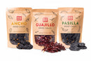 Three upright bags of Rico Rico Dried Mexican Chiles 3-Pack – Ancho, Guajillo & Pasilla are displayed, with a handful of authentic Mexican chiles scattered in front. The Rico Rico packaging showcases chili illustrations and heat level indicators.