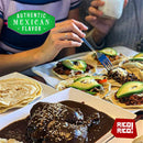 Two people enjoy tacos topped with avocado and shredded cheese, savoring the rich taste of authentic Mexican spices. A plate of tortillas and a dish of chicken in mole sauce highlight the bold flavors created using Rico Rico Chile Powder 6 Pack Bundle (24 oz Total) – featuring Ancho, Arbol, Chipotle, Guajillo, Habanero, and Pasilla chiles for genuine Mexican flavor. The image displays “Authentic Mexican Flavor” alongside the Rico Rico logo.