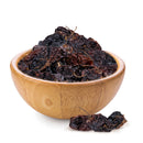 A wooden bowl filled with dried black raisins, with a few raisins and Rico Rico Chipotle Morita Dried Chiles—smoky and medium heat Mexican peppers—scattered on the white surface beside the bowl.