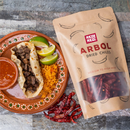 A plate featuring a beef taco, orange rice, lime wedges, and salsa sits beside a bag of Rico Rico Chile de Árbol Dried Chiles – Hot & Authentic Mexican Peppers, bringing the bold flavors of authentic Mexican cuisine to your table. Loose Rico Rico dried chiles are scattered nearby on a stone surface, highlighting the vivid color and distinctive character of these classic Chile de Árbol peppers.
