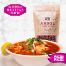 A bowl of vibrant red soup garnished with cilantro, diced onions, and fresh lime sits next to a bag labeled "Rico Rico Chile de Árbol Dried Chiles – Hot & Authentic Mexican Peppers," emphasizing the bold, authentic flavor of Chile de Árbol. Pink text reads: "Authentic Mexican Flavor" and "Rico Rico! Medium to High Heat.