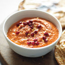 A white bowl of creamy red dip topped with pomegranate, chili flakes, and SALTOPIA BLT Salt by SALTOPIA Infused Sea Salts sits on a wooden surface beside flatbread.