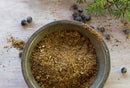 A metal bowl filled with ground seasoning sits on a wooden surface, surrounded by whole dried juniper berries and rosemary sprigs—perfect for creating an aromatic meat rub or wild game spice blend with the World Spice Rocky Mountain Rub from World Spice.