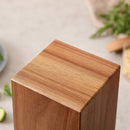 A close-up of the top corner of the Norpro Rotating Magnetic Knife Holder, featuring a smooth acacia wood block with natural grain, set on a light surface with blurred greenery and other objects in the background.