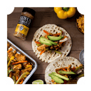 Two chicken fajitas on tortillas with avocado slices, next to a tray of seasoned chicken and vegetables, a lime wedge, Rufus Teague Chick N' Rub bottle, yellow bell pepper, and a bowl of rice.