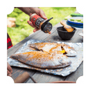 A person sprinkles Rufus Teague Fish Rub onto two whole fish on a foil-covered tray outdoors, with various utensils and bowls from Rufus Teague nearby on a wooden table.