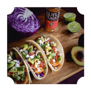 Three tacos filled with fish, diced tomatoes, avocado, cilantro, and cheese sit on a wooden board, surrounded by a halved avocado, purple cabbage, lime halves, and a bottle of Rufus Teague Fish Rub.