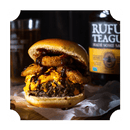 A juicy cheeseburger topped with onion rings and drizzled with Rufus Teague Honey Sweet BBQ Sauce sits on parchment paper, with a glass and a bottle of Rufus Teague sauce blurred in the background.