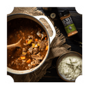 A pot of beef stew with carrots and celery sits on a rustic table next to mashed potatoes, a wooden spoon, and a bottle of Rufus Teague Meat Rub.