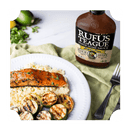 A plate of grilled salmon, cauliflower rice, and zucchini sits next to a fork, with a bottle of Rufus Teague Slim N' Sweet Sugar Free BBQ Sauce in the background.