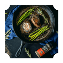 A cast iron skillet with two cooked steaks, asparagus, and rosemary sits on a blue cloth. Nearby, a bottle of Rufus Teague Steak Rub by Rufus Teague and fresh rosemary are placed on the dark surface.